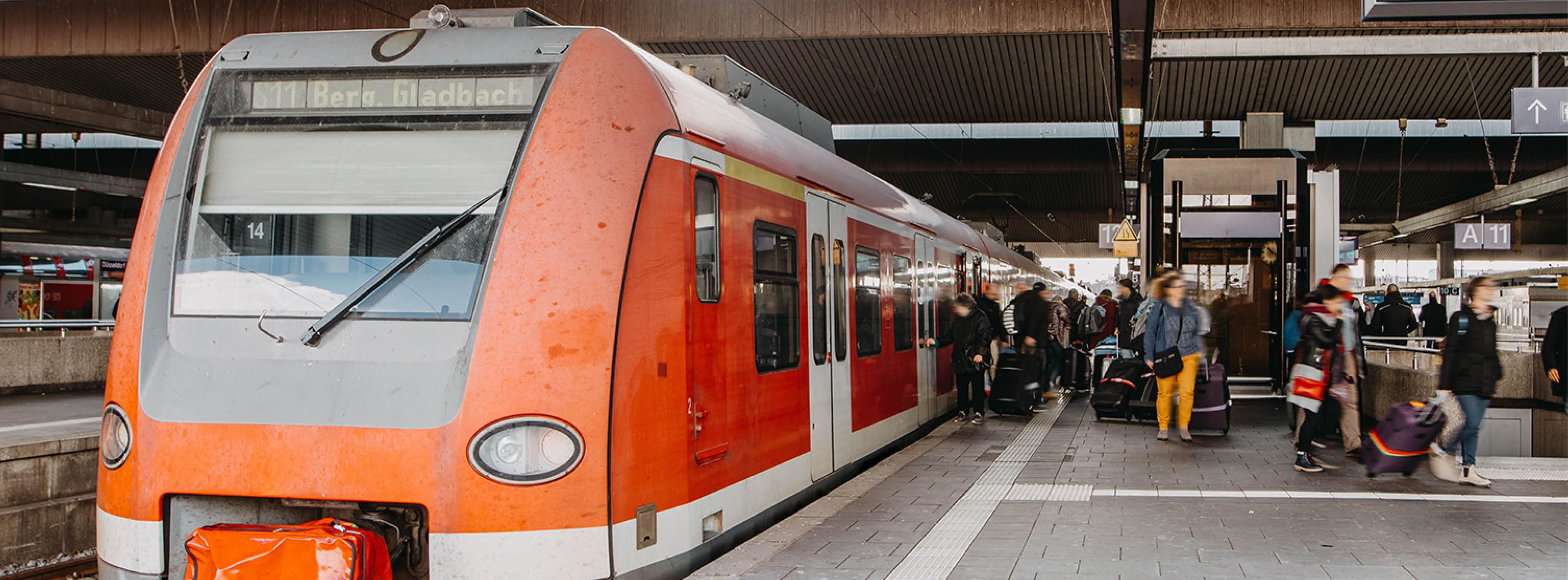 Ein Regionalzug der Linie S11 mit dem Ziel Bergisch Gladbach hält an einem Gleis eines Bahnhofs. Menschen steigen ein und aus.