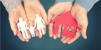 Zwei ausgestreckte Hände halten einen Scherenschnitt in Form von drei Personen. Daneben zwei weitere ausgestreckte offene Hände, die einen Scherenschnitt in Form eines Hauses halten. 