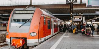 Ein Regionalzug der Linie S11 mit dem Ziel Bergisch Gladbach hält an einem Gleis eines Bahnhofs. Menschen steigen ein und aus.